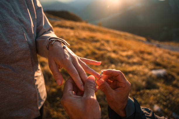 A mature gentleman tenderly clasps his partner's hand while placing an engagement ring during a heartfelt proposal amidst a mountainous outdoor setting.