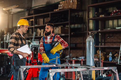 Colleagues in a work facility talking while making a production plan together. Both wearing sleeved plaid shirts and work overalls as well as safety gear. Dangerous work environment. Copy space.