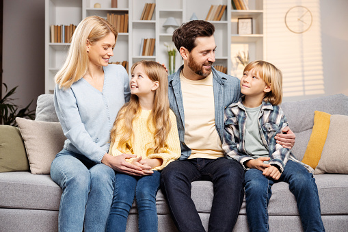 Loving family at home theme: Seated snugly on soft settee in heart of their living space, mother, father, daughter, son share laughter and affectionate cuddles, symbolizing perfect home life concept
