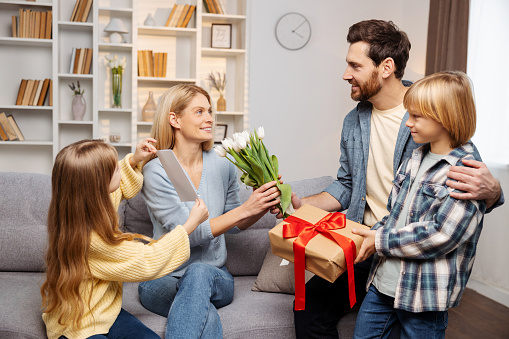 Celebrating Mother's Day indoors: Eager son and daughter give a handcrafted card and boxed gift to their thrilled mother on the snug sofa, with dad offering a gorgeous bunch of flowers