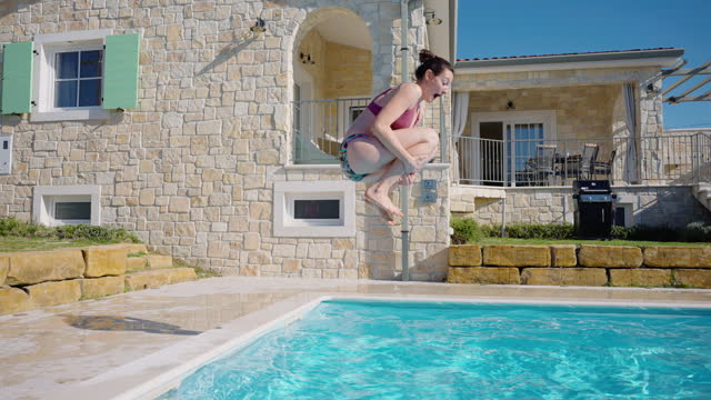 SLO MO Young Woman in Bikini Jumping with Hugging Knees into Swimming Pool at Holiday Villa on Sunny Day