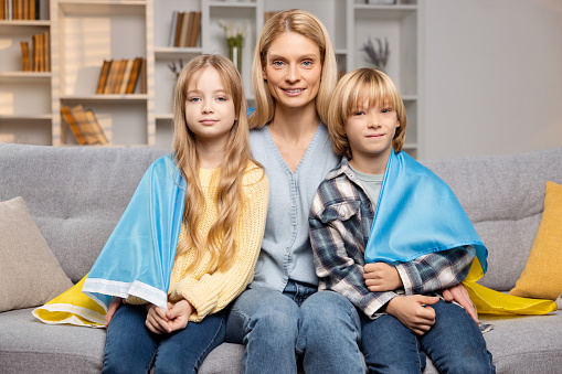 Hermosa madre abrazando a sus hijos bajo la bandera ucraniana, sentados juntos en el sofá de la sala de estar photo