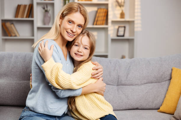istock Smiling mother hugging her little daughter. Both sitting on couch, looking at the camera 2064388080