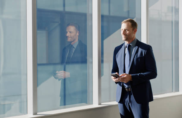 istock Window, thinking and businessman with smartphone in office building for planning, vision or inspiration. Workplace, idea and professional person with technology for networking, communication or break 2064414423