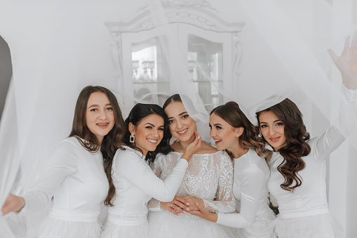 Young bridesmaids in white silk dresses hug and rejoice in the bride's room. Beautiful women celebrating bachelorette party standing in room.