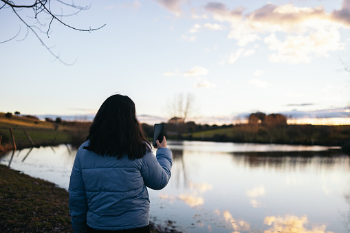 Rear view of a young woman in a down coat watching the sunset on a lake in nature and taking photos with the smartphone