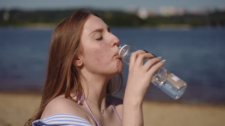 A girl drinks water from a plastic bottle while sitting on a sandy beach near a river.