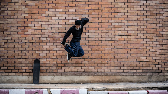 A cool, energetic Asian man in fashionable clothes is jumping outdoors, practicing his breakdance on the street.
