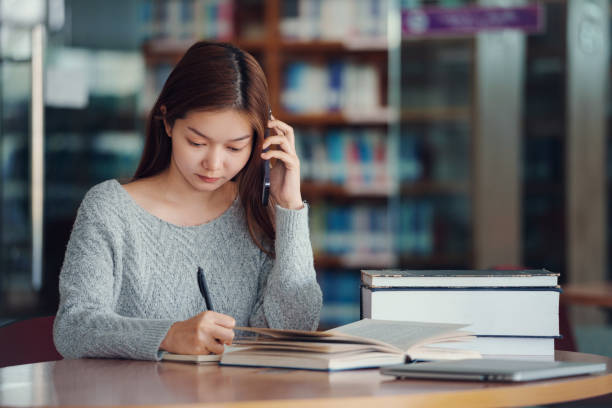istock Unhappy young asian woman talking on mobile phone while reading book in library. 2064739557