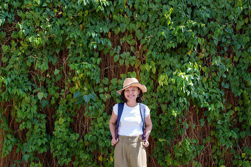 A happy solo Asian woman traveler, sporting a straw hat and backpack, poses against a leafy wall while exploring Georgetown, Penang.