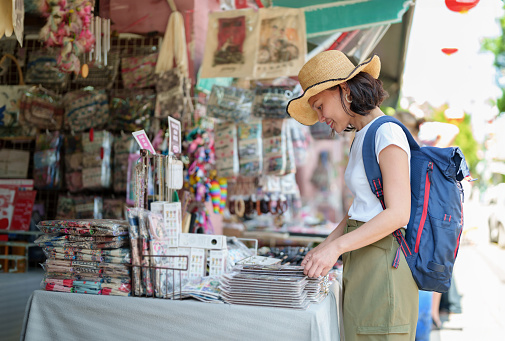 A solo Asian woman traveler stop at a street stall adorned with local crafts and souvenirs. She carefully examines each item, considering which ones will serve as cherished mementos of her journey.