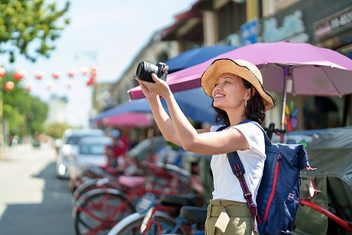 A cheerful Asian woman traveler, wearing a straw hat, takes photos with her camera while exploring Georgetown, Penang. She travels light, capturing the lively streets and colorful buildings. It's a simple, joyful way for her to explore and make memories.