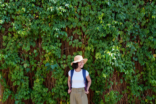 A happy solo Asian woman traveler, sporting a straw hat and backpack, poses against a leafy wall while exploring Georgetown, Penang.
