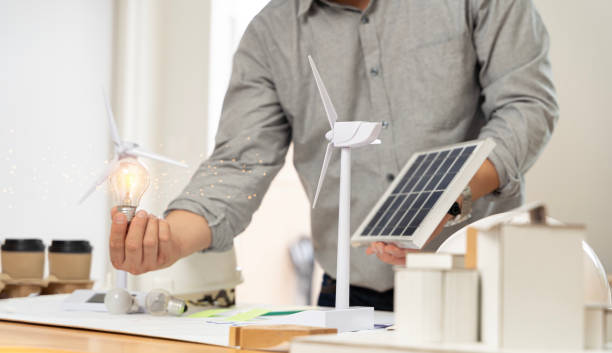 istock Close-up at a light bulb, An engineer sits holding a light bulb and model of wind turbine. with solar panels and model house on the table. 2064835537