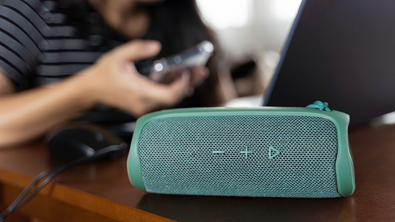 Close-up of a stylish green Bluetooth speaker placed on a table at home. The woman in the background sits at a table and works using a laptop and a smartphone on the table next to her.