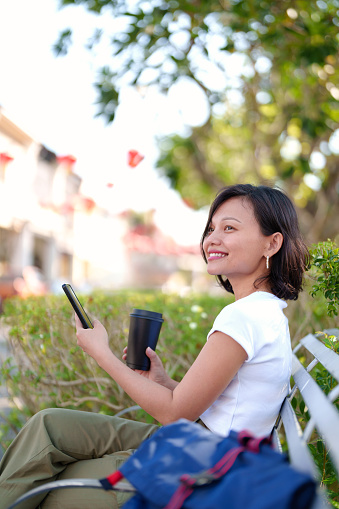 A young Asian woman solo traveler takes a coffee break in an urban park in the city. She sits amidst the greenery, using her smartphone and enjoying a cup of coffee, taking a moment to relax during her vacation.