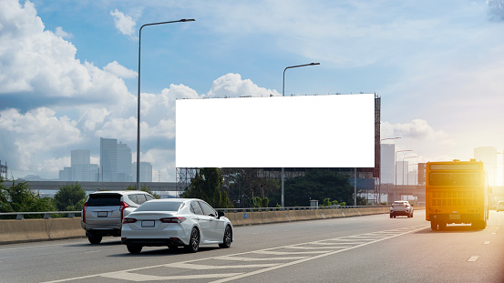 A large, blank billboard on the side of a highway. The billboard is white. It is located in a rural area, with trees and grass in the background. The sun is shining and the sky is blue, clipping path.