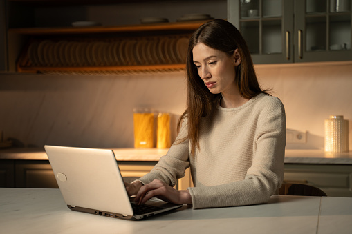 A focused young woman is seated at a kitchen counter, engaged in work or study on her laptop, with soft lighting illuminating her from above. Woman concentrates on her task.
