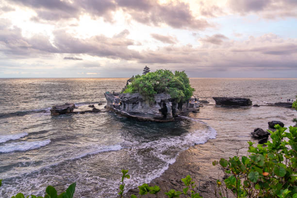 Tanah Lot temple on cliff in Indian ocean. Waves are breaking on a rock cliff off the coast of Bali. Hindu temple built on a rock in the sea. Lush tropical vegetation on top of a cliff.