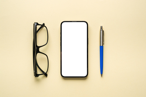 Top view of glasses, ballpoint pen and smartphone with blank screen on yellow background