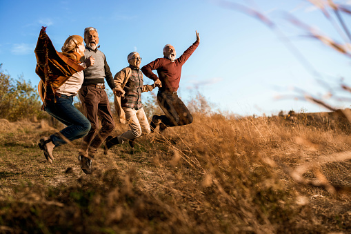 Happy senior couples having fun while holding hands and jumping in a meadow in autumn day. Copy space.