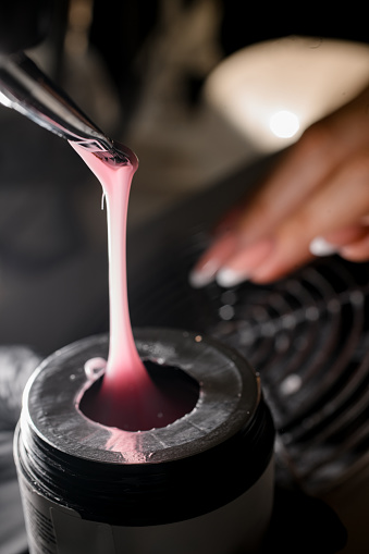 Manicurist's hands pick up pink gel from a jar on a brush to apply it to the client's nails