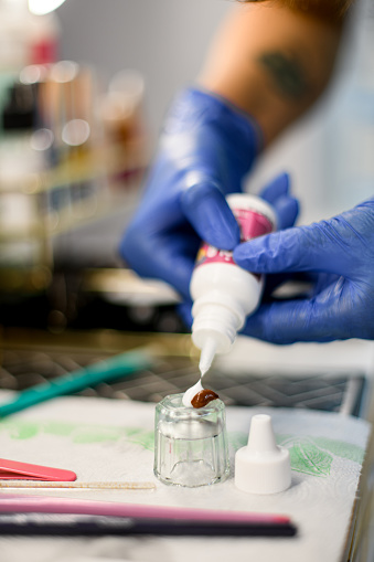 Female hands in blue gloves squeezing paint and oxidizer into a glass cup for mixing eyebrow paint on white table