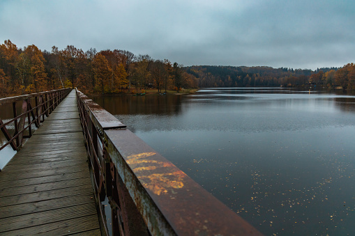 Beautiful view to long steel and wood bridge over big and silent lake with autumn golden trees and bushes around at cloudy and rainy morning