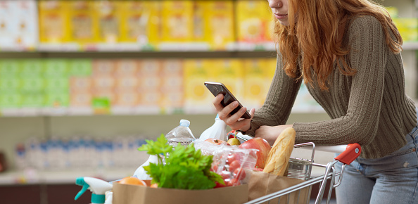 Young woman doing grocery shopping at the supermarket, she is pushing a full shopping cart and using her smartphone