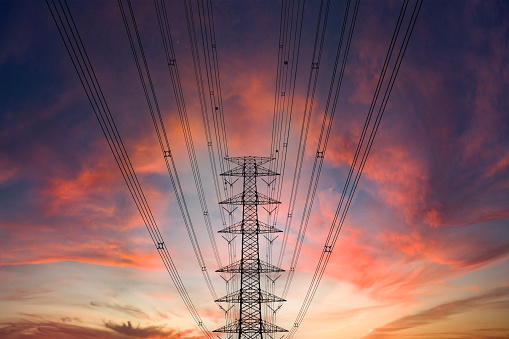 A power line crosses the foreground of a dramatic sunset. The setting sun casts a warm glow over the landscape, silhouetting the power line and its supports. High voltage wires concept.