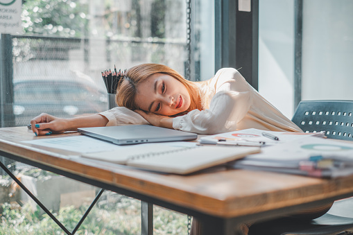 Overcome by exhaustion, a young woman rests her head on her desk amidst a spread of work materials, a moment of stillness in her busy day captured in a light-filled office space.