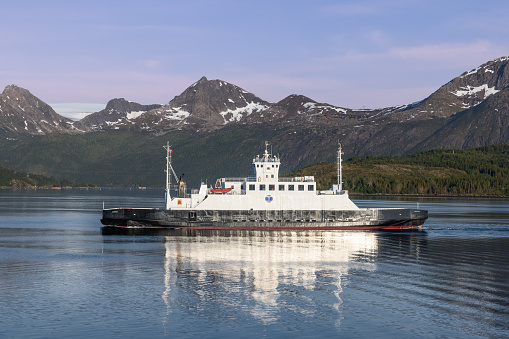 Meloy - Norway. June 23, 2023: The ferry 'Narvik' embarks on its journey across a placid northern fjord, reflecting on the mirror-like surface with a backdrop of rugged peaks