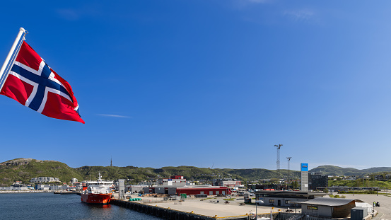 Bodo - Norway. June 17, 2023: A vibrant Norwegian flag in the photo's left corner flutters against a summer blue sky, overlooking the bustling tourist and trade port of Bodo as a gateway to the Lofoten Islands