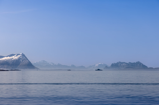 Bodo - Norway. June 17, 2023: View from a ferry headed to Lofoten Island, featuring the North Sea's hushed waters, snow-dusted, contoured mountains, and a military ship cruising along the coast