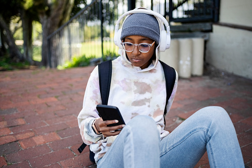 Teen african american girl wearing wireless headphones listening to songs on smartphone while sitting outside on paved road