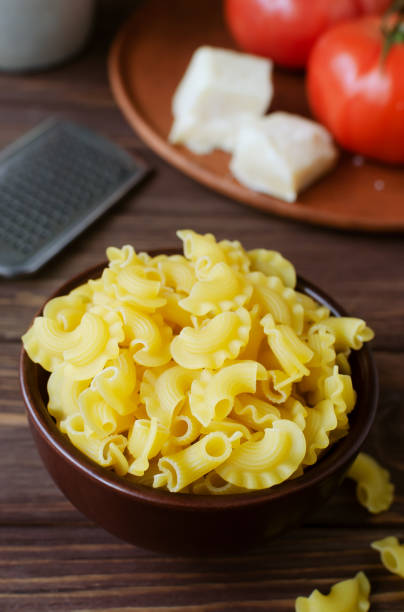 Raw Creste Di Gallo pasta in a ceramic bowl with fresh tomatoes and parmesan cheese on a brown wooden board. Rooster comb. The concept of traditional Italian cuisine. Vertical orientation. stock photo