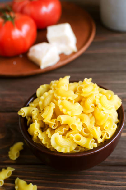 Raw Creste Di Gallo pasta in a ceramic bowl with fresh tomatoes and parmesan cheese on a brown wooden board. Rooster comb. The concept of traditional Italian cuisine. Vertical orientation. stock photo