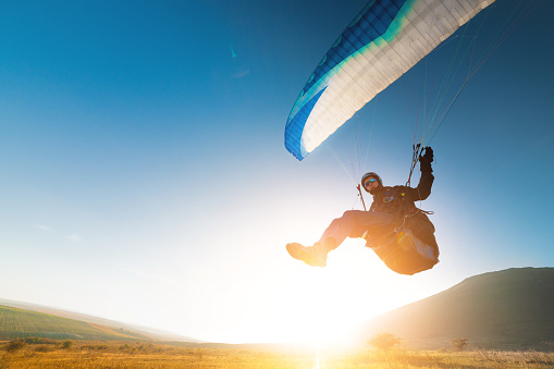 A paraglider takes off from a mountainside with a blue and white canopy and the sun behind. A paraglider is a silhouette. The glider is sharp, with little wing movement. A male paraglider launches a paraglider into the air.