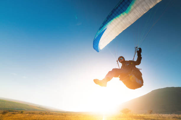 A paraglider takes off from a mountainside with a blue and white canopy and the sun behind. A paraglider is a silhouette. The glider is sharp, with little wing movement. stock photo