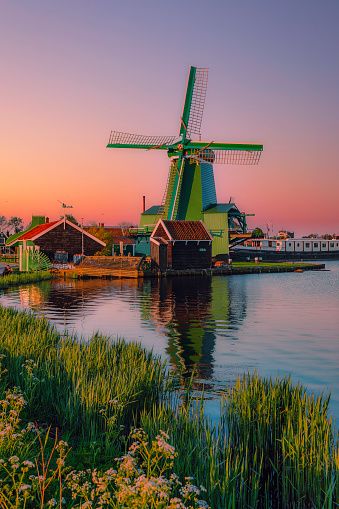 View traditional Dutch windmills along the canal in Zaanse Schans, Zaandam, Netherlands, with fishing shelters, boats, and shipyard equipment. It is a usual gray mill with orange wings near the water. The rural scene at the famous tourist site in Holland shows windmills at sunrise or sunset with a dramatic sky