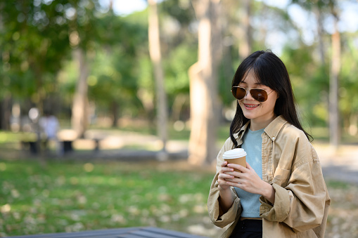 Close-up portrait young pretty woman drinking coffee from black paper cup sitting on a bridge in the park.