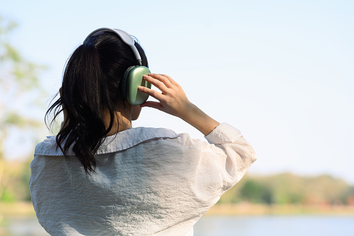 Rear view of relaxed young woman standing by the lake and listening music with headphones.