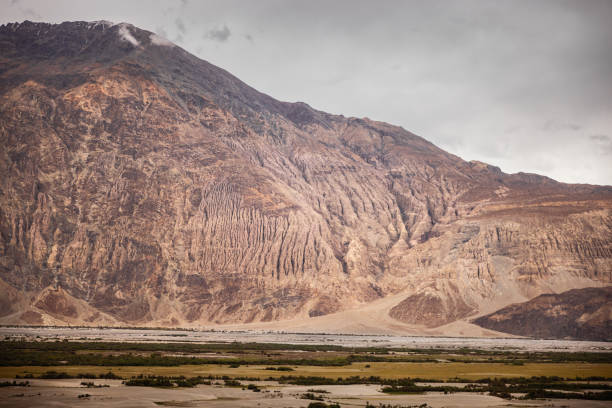 Padmasambhava statue placed in a vast field with majestic mountains in the backdrop, under a sky filled with clouds and surrounded by greenery like grass, trees, and natural landscape, Zanskar, Himalayas