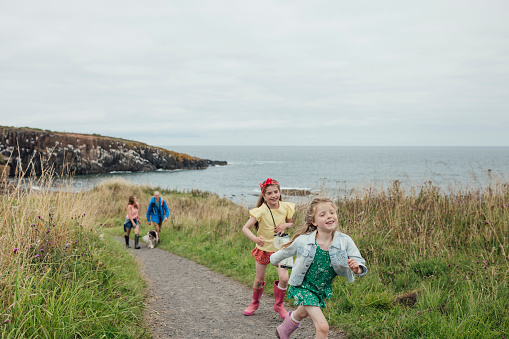 A wide full-length shot of a family enjoying a day out at the coast in Northumberland, North East England. Two sisters run ahead smiling, close to the camera, whilst their parents walk further behind with the pet dog. They are all wearing casual clothing on an overcast summer's day. The path slopes away from the camera and is lined with lush grass and foliage. The sea and a rocky headland are visible in the background.