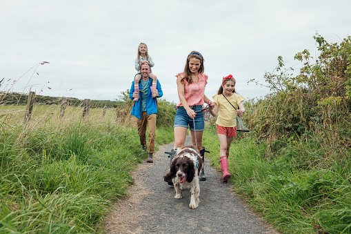 A wide full-length shot of a family of four walking along a coastal path in Northumberland, North East England. They are all smiling, wearing casual clothing on an overcast summer's day. The path is lined with lush grass and foliage. The female is leading their pet dog, holding her daughter's hand, whilst the male carries their other daughter on his shoulders.