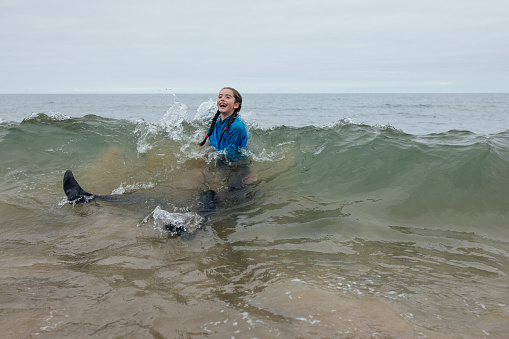 A shot of a father and his daughter playing in the waves at a beach in Northumberland, North East England. The father's face is completely obscured as his daughter playfully pushes him underneath the surface of the water. The young girl is smiling joyfully, they are wearing wetsuits and rash guards, and it is an overcast summer's day.