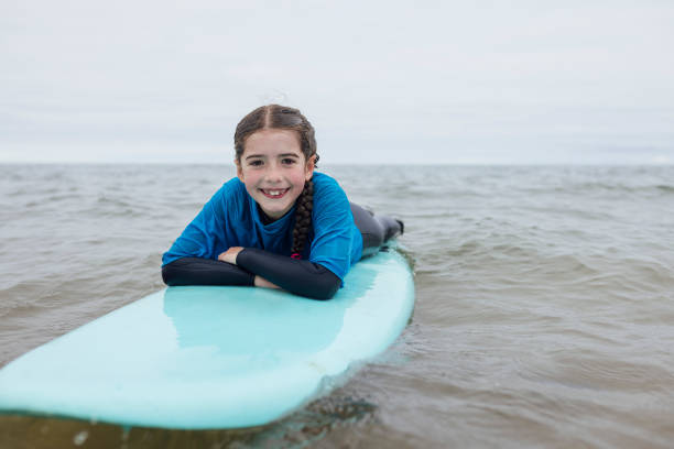 A shot of a young girl lying on a surfboard, leaning on her crossed arms, in the sea in Northumberland, North East England. She is smiling directly at the camera, wearing a wetsuit and rash guard. She has vitiligo which is partially visible on her hand. She has lost some primary teeth to make way for her permanent teeth.