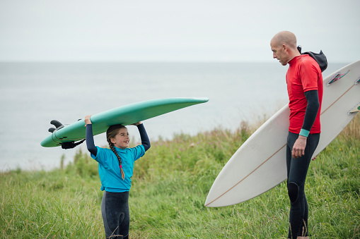 A side-view shot of a father standing with his daughter who is balancing her surfboard on her head in the sand dunes before going surfing at a beach in Northumberland, North East England. She is smiling up at her father, both wearing wetsuits and rash guards. It is an overcast summer's day.