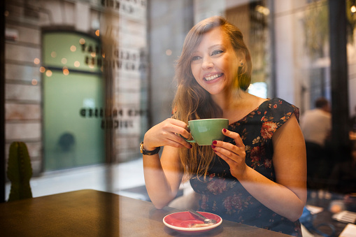 Portrait of beautiful woman drinking coffee, wears floral dress, broadly smiles sitting inside bar at city.
