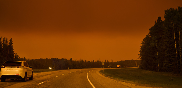 Cars driving on a highway under a sky glowing orange from a forest fire burning in the countryside